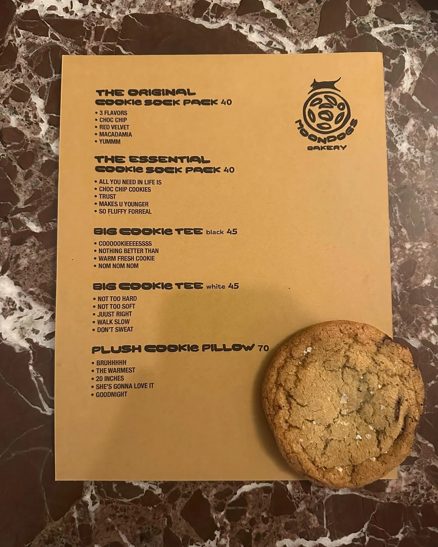 The printed menu card on a marble counter with a cookie set on it. Product names in the painted face, descriptions in a plain condensed sans.